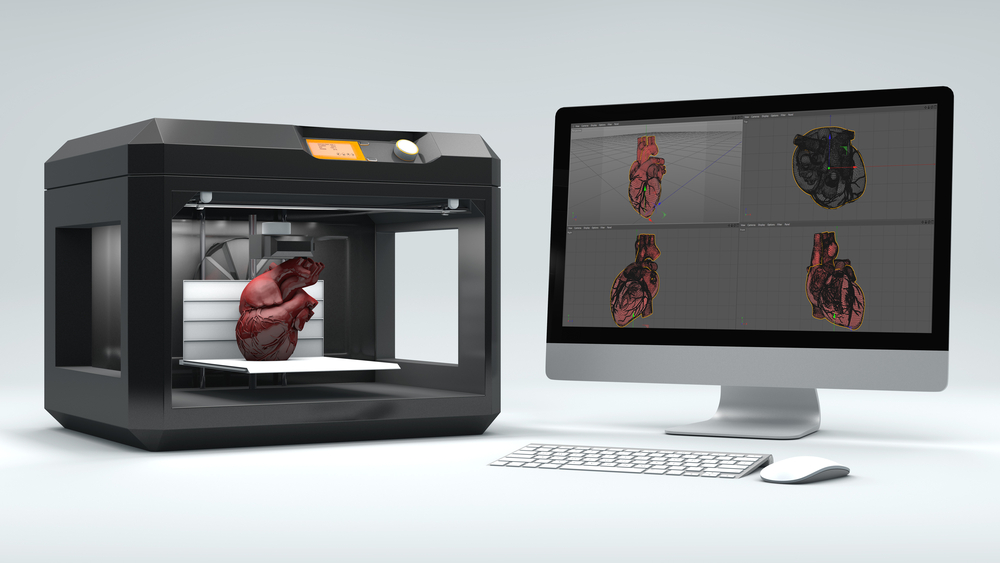 An example of a 3D printed heart inside a 3-D printing device next to a desktop computer displaying the human heart.