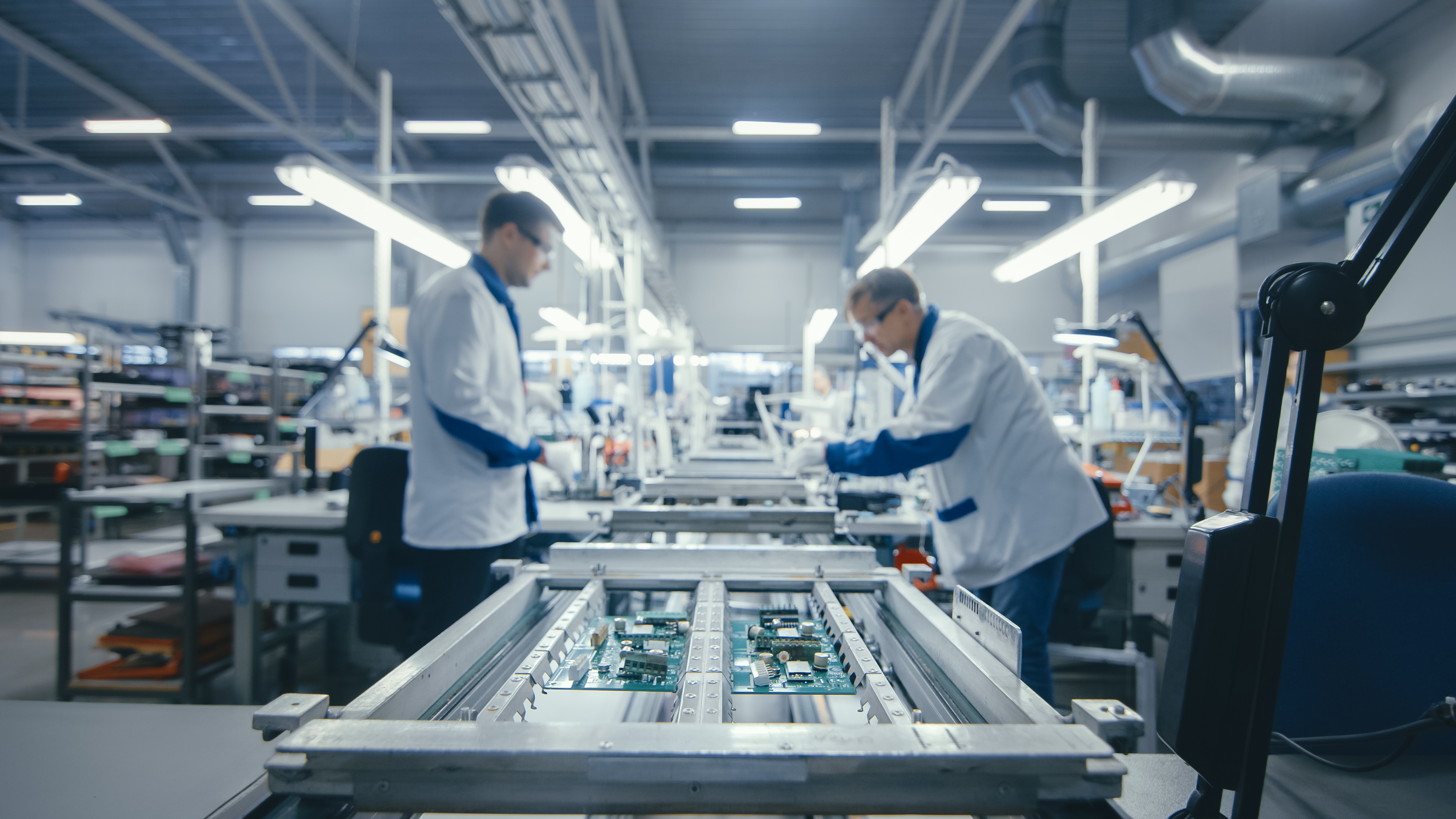 Technicians in personal protective equipment inspect circuit boards by hand on an assembly line in a high-tech facility.