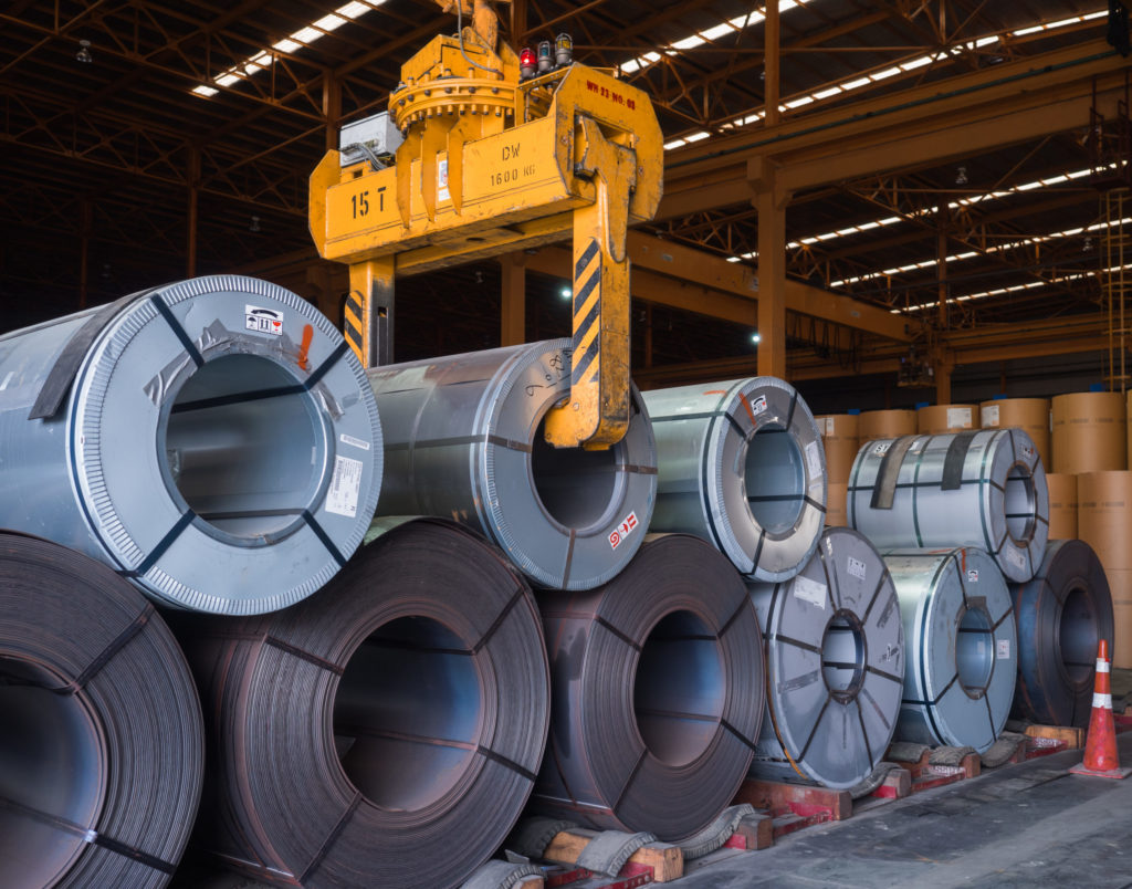 An overhead crane picks up a spool of sheet metal.