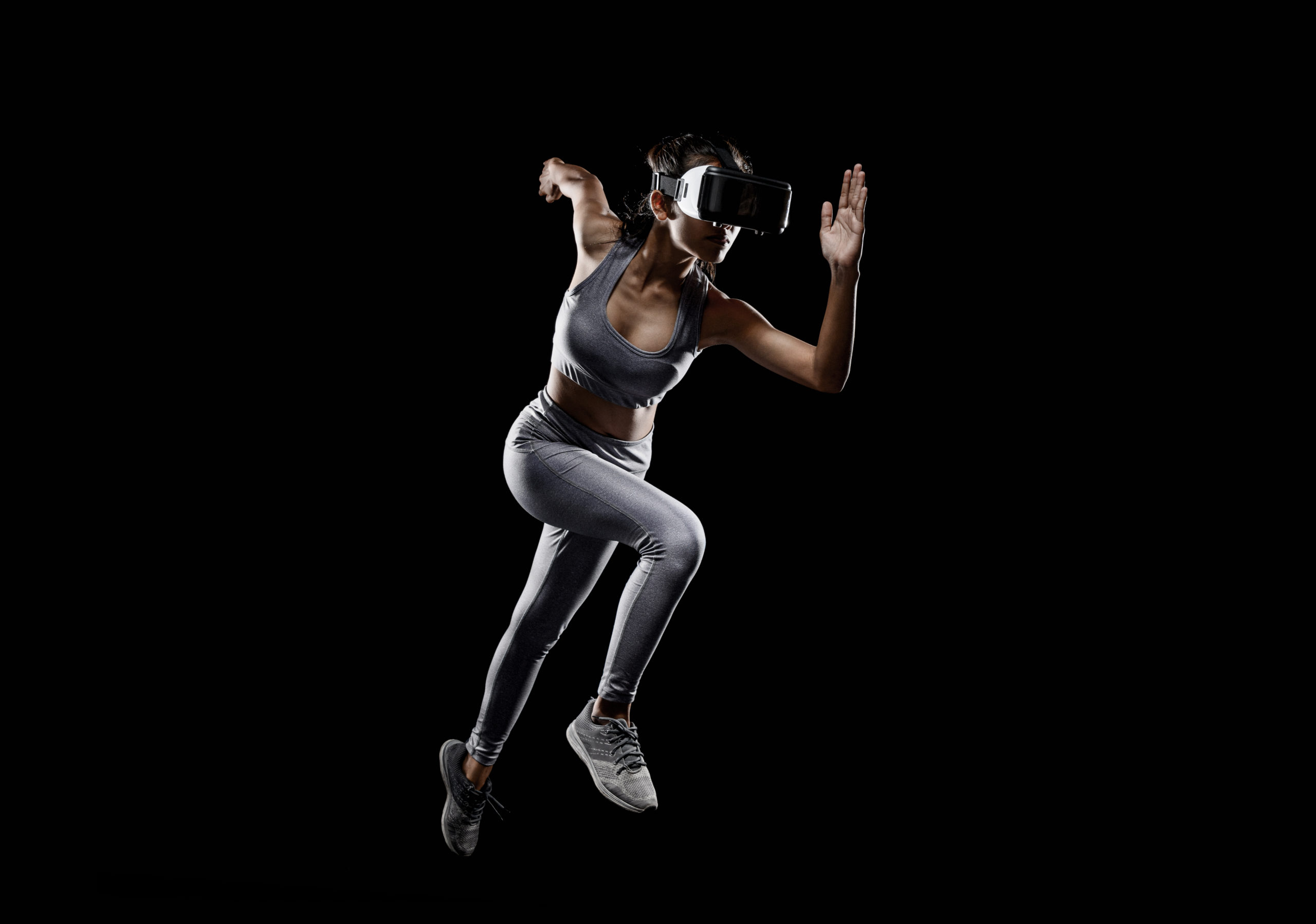 Woman in athletic clothing and VR goggles running intensely. Black background.