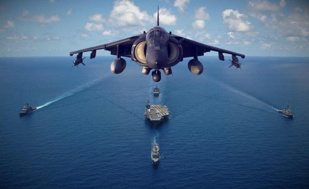 A harrier jet in flight over an aircraft carrier and accompanying ships.